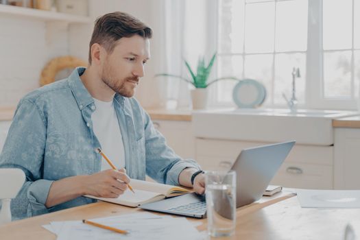 Concentrated young businessman writing notes on agenda and working remotely on laptop in internet from home while sitting at kitchen table. Freelance and work online concept