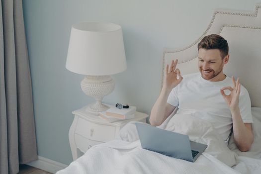 Happy positive young man sitting in cozy bed after waking up in early morning, chatting by video call on laptop and smiling agrees with friend joining fingers in ok sign with both hands