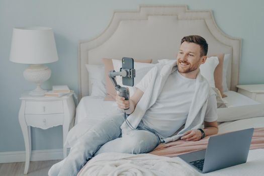Young bearded man video chatting with someone close to him, holding phone with gimbal, happy to be talking, lying on comfortable bed with big headboard, open notebook in front