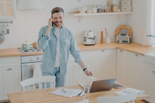 Handsome smiling bearded man wearing casual clothes working on laptop computer while standing at kitchen table at home and talking on mobile phone with friend. Freelance and distance work concept