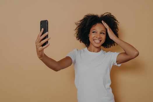 Happy charming dark skinned female 20s in white tshirt making photo selfie on modern smartphone and smiling cheerfuly, isolated over beige wall. Positive young african woman taking photo on cellphone