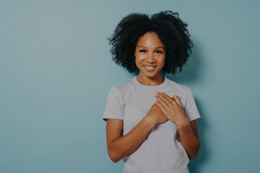 Grateful hopeful happy black woman holding hands on chest, satisfied and sincere african lady expressing love from heart, isolated on blue studio background with copy space. Positive emotions concept