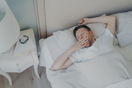 Young sleepy man waking up in morning after night sleep while lying in bed, yawning and covering his mouth with hand and keeping eyes closed. Sleeping and bedtime concept
