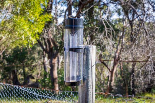 A rain gauge bolted to a wooden fence posts near trees