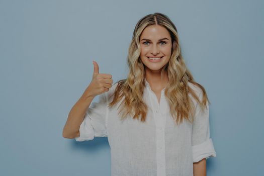 Cheerful blonde girl in white shirt showing thumbs up gesture with her hand while smiling, admitting that she liked that, standing alone next to blue wall. Positive feedback concept