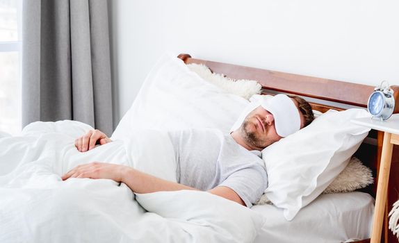 Man wearing pajamas and eye sleeping mask napping in the bed in the morning time. Guy resting in the room with daylight. Male person under blanket at home