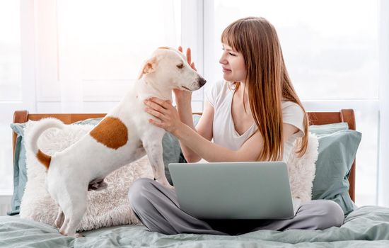 Attractive girl sitting in the bed with grey laptop and petting her cute dog. Young beautiful woman with pet and notebook in morning time in the bedroom. Concept of remote work from home