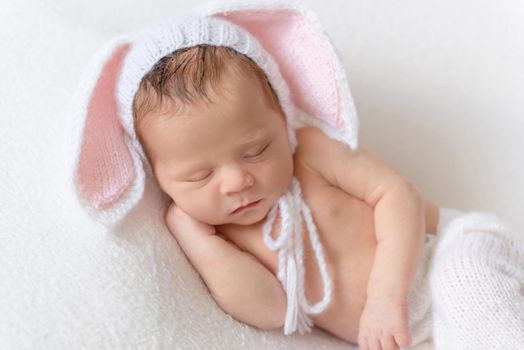 Little cute baby in white knitted pants holding white toy bunny and sleeping on white soft bedcover