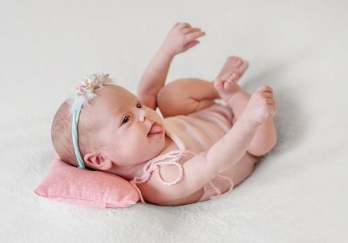 Little smiling baby in pink knitted suit joyfully lying on pink dotted pillow and white soft blanket