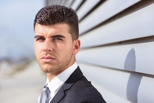 Young businessman near a modern office building wearing black suit and tie. Man with blue eyes