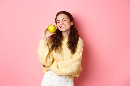 Healthcare, stomatology and people concept. Happy girl show her white perfect teeth, smile and green apple, eating healthy food and fruits, standing against pink background.