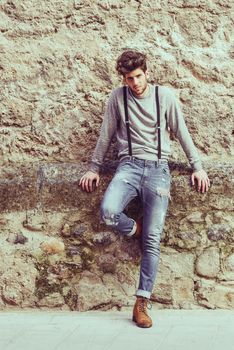 Portrait of young man wearing suspenders and blue jeans in urban background with modern haircut