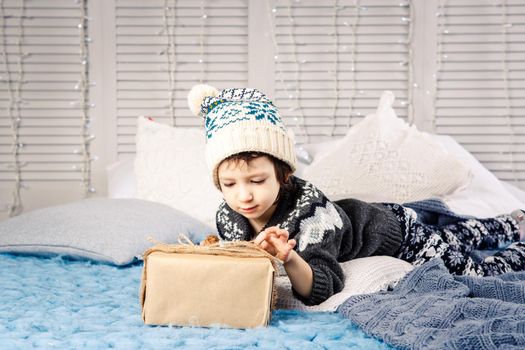 little girl the child sitting in pajamas and hat on the bed with garland of light bulbs with gifts boxes wrapped in a non-colored paper decorated with cones on blue knitted coverlet.Christmas concept.