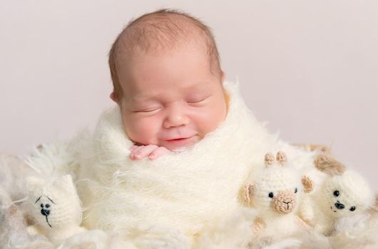 Portrait of smiling gorgeous baby asleep in soft beige wrap with plush toys around