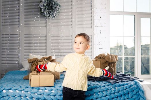 little funny playful boy a child sits on a bed on Christmas day with gift boxes in white wool knitted sweater and big bright mittens on it and laughs out loud. In interior there is a festive decor.