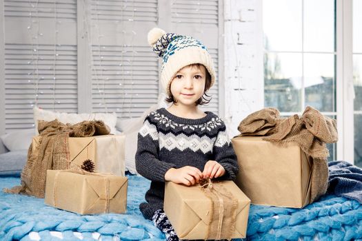 little girl the child sitting in pajamas and hat on the bed with garland of light bulbs with gifts boxes wrapped in a non-colored paper decorated with cones on blue knitted coverlet.Christmas concept.