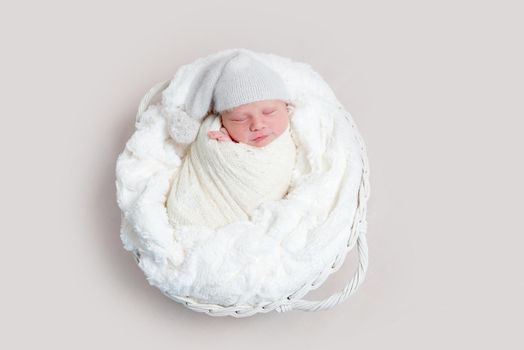 Top view of newborn baby wrapped in white wrap wearing white bonnet and lying on white round basket with soft blanket. Newborn baby in white outfit