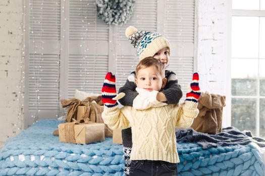 Christmas concept.Children brother and sister in embrace of the house in the bedroom near bed with boxes,gifts on back of the decor and windows on sunny day.Wear a warm woolen garment cap and mittens.