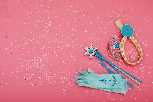 From above of magic wand and gloves placed near crown and pigtail on pink background with silver glitter
