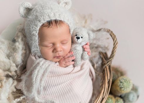 Little baby in cute white knitted beanie with ears and covered in light pink coverlet sweetly sleeping in the basket with cactus nearby