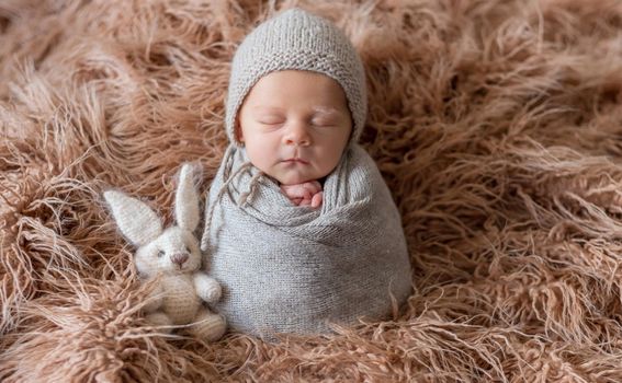 Little cute baby weared in gray knitted beanie and covered with gray knitted blanket sweetly sleeping with bunny nearby on the soft light brown bedcover