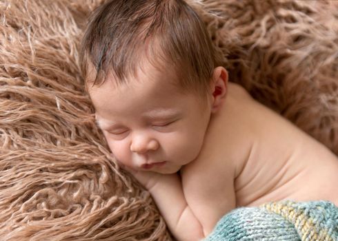 Little baby weared in light blue-yellow knitted beanie and pants sweetly sleeping on the light brown soft coverlet