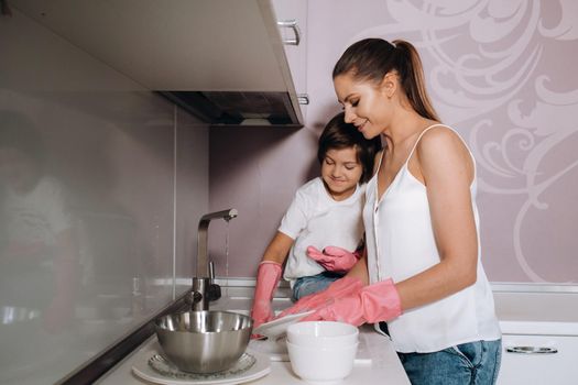 housewife mom in pink gloves washes dishes with her son by hand in the sink with detergent. A girl in white and a child with a cast cleans the house and washes dishes in homemade pink gloves.A child with a cast washes dishes and smiles.