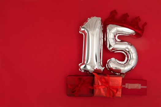 Foil silver air balloons showing number 15 fifteen placed on red background with wrapped presents for concept of birthday anniversary