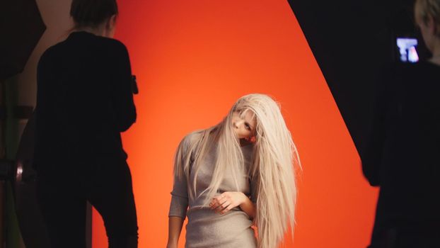 Fashion backstage: blonde girl model plays long hair - photographer take a picture in studio, red background