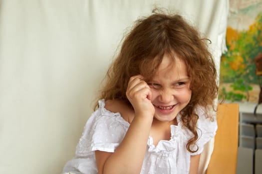Happy little girl touching curly hair and looking away with smile while sitting near curtain