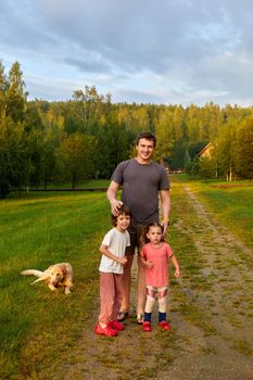 Adult man with daughter and son standing on path near loyal dog on summer weekend day in nature