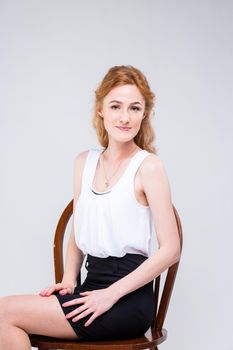 Portrait of beautiful business woman with long red, curly hair sitting on wooden chair on white background in studio. Dressed in a white blouse with a short sleeve and black short skirt and high heels