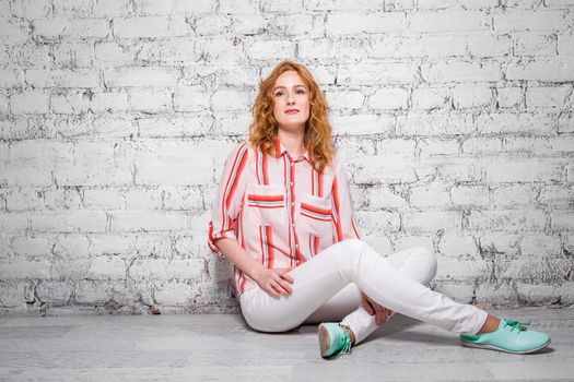 Young beautiful caucasian woman student is sitting on the floor near a white brick wall. Girl with red long curly hair with freckles on her face in bright summer clothes.