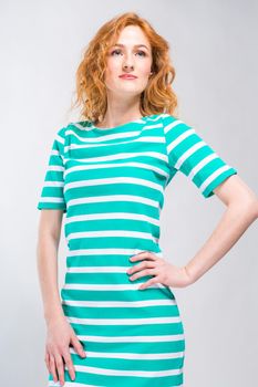 A lap portrait of a young, beautiful woman with red curly hair in a summer dress with stripes of blue in the studio on a gray background. Theme of summer vacation, tourism and summer clothes.