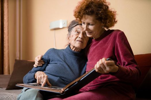 Positive aged ladies looking album photos sitting sofa at home, cheerful friends. Senior woman and her mature nurse watching photo album. Granny showing her daughter memories from the past.