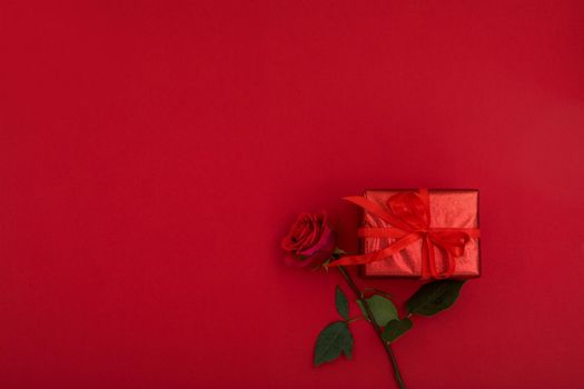 Top view of small present box wrapped in red paper with ribbon placed near blooming rose on bright red background with blank space for romantic