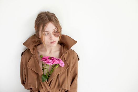 Young female model with dark spot under eye dressed in brown coat with bright blooming flowers standing against white wall