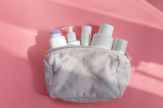 From above of assorted white bottles of skin care products placed in cosmetic bag on pink background lit by sunlight
