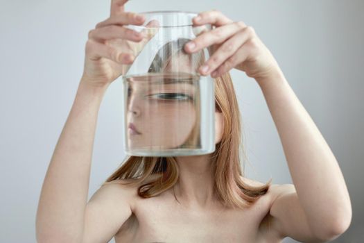 Unrecognizable female with bare shoulders looking at camera through transparent glass jar filled with water