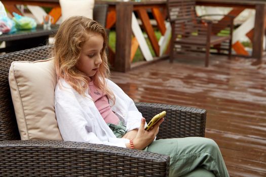 Preteen girl in casual clothes leaning on pillow and watching video on cellphone while sitting on wicker armchair on terrace