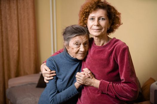 Elderly old cute woman with Alzheimer's very happy and smiling when eldest daughter hugs and takes care of her, at home on sofa. Theme aging and parenting, family relationships and social care oldest.
