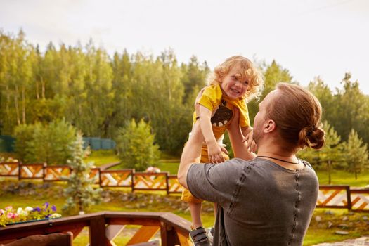 Unrecognizable father lifting adorable son with curly hair standing on wooden railing in rural area against forest with green trees