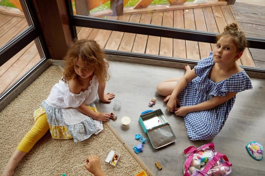 From above sisters in casual clothes sitting on floor near window and playing together with toys on weekend day at home