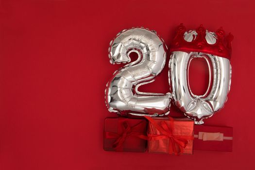 Foil silver air balloons showing number 20 twenty placed on red background with wrapped presents for concept of birthday anniversary