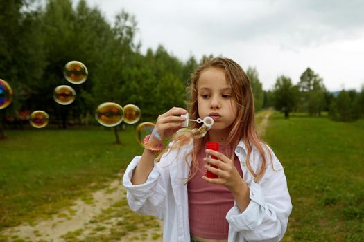 Cute girl in casual clothes blowing soap bubbles while standing on path on overcast summer day in nature
