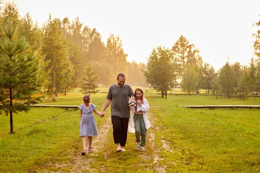 Content loving father walking with preteen daughters along green meadow in village in sunset