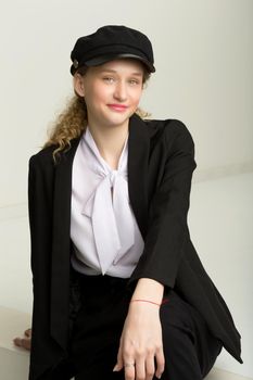 Portrait of cheerful girl in elegant suit and cap. Charming stylish young woman in fashionable black blazer, white blouse and cap sitting relaxed against gray background in studio