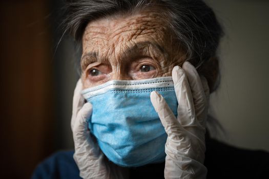 Portrait senior woman holding head hands, sad worried emotion in protective medical mask, gloves against virus and infection at home. Old gray hair female quarantine stay at home, loneliness and fear.