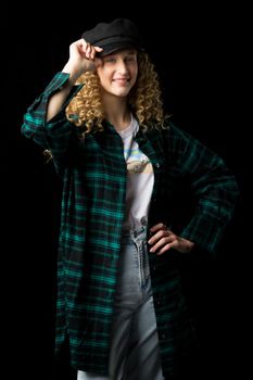 Beautiful blonde girl touching her stylish black cap. Portrait of girl with wavy hairstyle wearing plaid cotton shirt and white t-shirt looking seriously at camera against black background