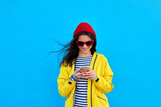 Cheerful young female in yellow jacket and in red hat browsing mobile phone on vivid blue background